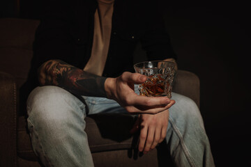 Young man with tattoos and glass of whiskey sitting in armchair on dark background, closeup