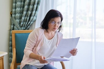Serious mature woman reading papers documents sitting in armchair at home.