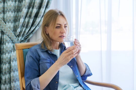 Sad Mature Woman Holding A Glass Of Water While Sitting In A Chair At Home.