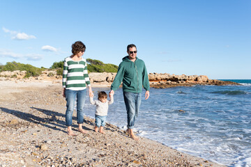 Young family having a walk at a mediterranean beach on a winter day. Father and mother supporting her little girl on her first steps. 