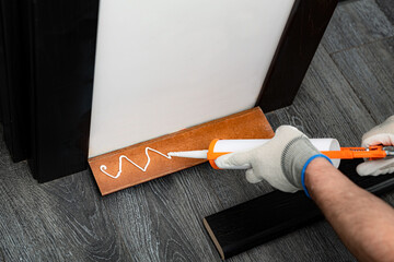 A handyman is putting a silicon glue from the tube on a wooden baseboard before attaching it to the wall - obrazy, fototapety, plakaty