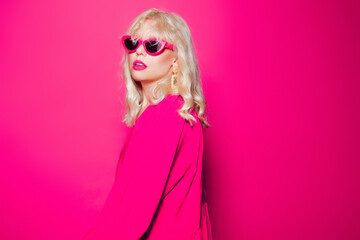 The blonde poses on a pink background in stylish pink glasses and a dress. Girl model posing,...