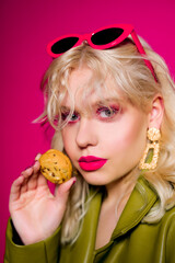 Obraz premium The blonde is eating cookies. High fashion, woman in earrings, gold jewelry. Bright pink glitter makeup. Pink glasses and stylish green cape