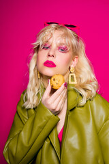 The blonde is eating cookies. High fashion, woman in earrings, gold jewelry. Bright pink glitter makeup. Pink glasses and stylish green cape