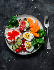 Breakfast, snack, tapas plate - sandwiches with red caviar, salmon, cream cheese, vegetables, spinach, boiled egg on a dark background, top view