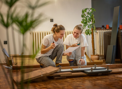 Two Young Women Girlfriends Or Lesbian Couple Assembling Furniture In Their New Home. Two Women Read The Cabinet Assembly Instructions.