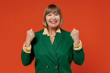 Elderly caucasian woman 50s wear green classic suit doing winner gesture celebrate clenching fists say yes isolated on plain orange color background studio portrait. People business lifestyle concept.