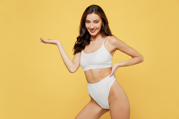 Fototapeta na wymiar Smiling lovely attractive young brunette woman 20s in white underwear with perfect fit body standing posing hold show empty arm with workspace area isolated on plain yellow background studio portrait