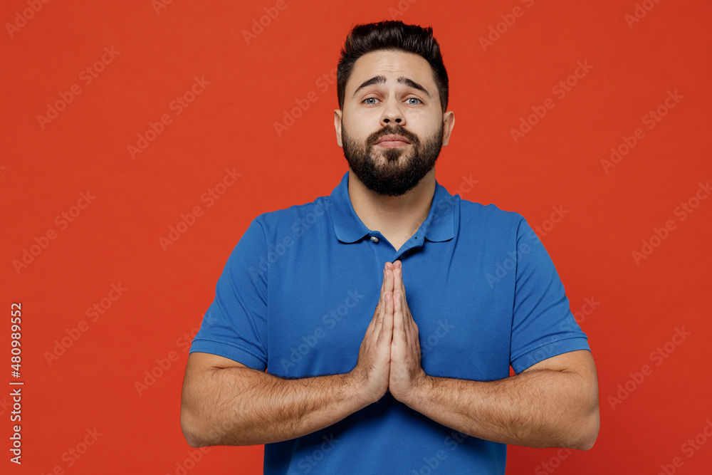 Sticker Young pleaded sad caucasian man 20s wear basic blue t-shirt hold hands folded in prayer gesture, begging about something isolated on plain orange background studio portrait. People lifestyle concept.