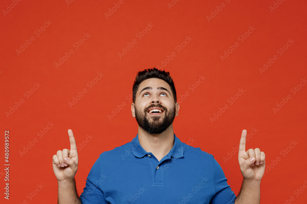 Poster Young happy smiling man 20s wear basic blue t-shirt looking camera point index finger overhead on workspace area mock up isolated on plain orange background studio portrait. People lifestyle concept.