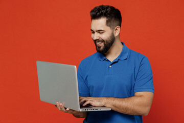 Young smiling happy man 20s in basic blue t-shirt hold use work on laptop pc computer browsing...