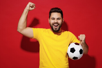 Excited happy young bearded man football fan in yellow t-shirt cheer up support favorite team hold soccer ball celebrate clenching fists say yes isolated on plain dark red background studio portrait.
