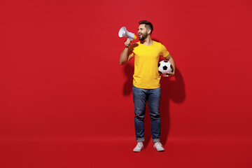 Fototapeta na wymiar Full size body length young bearded man football fan in yellow t-shirt cheer up support favorite team hold soccer ball scream aside in megaphone isolated on plain dark red background studio portrait.