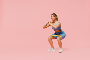 Full body young sporty athletic fitness trainer instructor woman wear blue tracksuit spend time in home gym use fitness rubber bands do squats isolated on plain pink background. Workout sport concept.