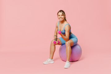 Full body young happy strong athletic fitness trainer instructor woman wear blue tracksuit spend time in home gym sit on fitball isolated on pastel plain light pink background Workout sport concept.