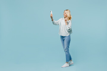 Full body elderly woman 50s wearing casual striped shirt doing selfie shot on mobile cell phone post photo on social network showv-sign gesture isolated on plain pastel light blue color background - Powered by Adobe