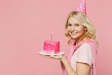 Side profile view elderly smiling cool happy woman 50s wear t-shirt birthday hat hold cake with candle look camera isolated on plain pastel pink background studio Celebration party holiday concept