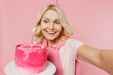 Close up elderly happy fun woman 50s in t-shirt birthday hat hold cake with candle do selfie shot pov on mobile phone isolated on plain pastel pink background studio Celebration party holiday concept