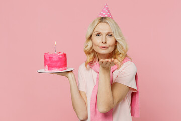 Obraz na płótnie Canvas Elderly happy satisfied caucasian woman 50s wear t-shirt birthday hat hold cake look camera blow air kiss isolated on plain pastel pink background studio portrait. Celebrating party holiday concept.