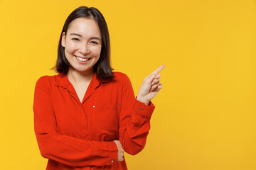 Naklejka premium Charming smiling young woman of Asian ethnicity 20s years old wears orange shirt looking camera pointing aside on workspace area copy space mock up isolated on plain yellow background studio portrait.
