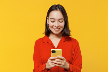 Charismatic vivid fun young woman of Asian ethnicity 20s years old wears orange shirt hold in hand use mobile cell phone reading searching chatting isolated on plain yellow background studio portrait. - Powered by Adobe