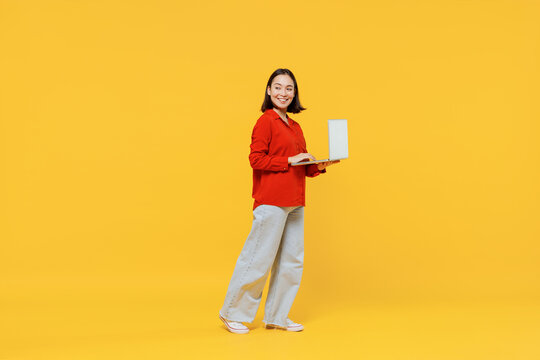 Full Size Body Length Vivid Smiling Happy Young Woman Of Asian Ethnicity 20s Years Old In Casual Clothes Look Back Hold Laptop Pc Computer Go Move Isolated On Plain Yellow Background Studio Portrait.