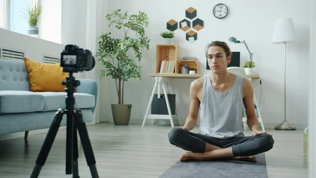 Young mixed race man is recording video tutorial about yoga using camera on tripod at home. Vlogging and active lifestyle concept.