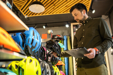 Bike shop manager checks helmet price information on tablet, seller makes an inventory in sports shop. Theme of small business selling bicycles. Seller in hands document checklist in bicycle store