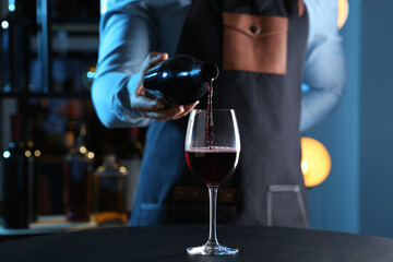 Bartender pouring red wine from bottle into glass at table indoors, closeup - obrazy, fototapety, plakaty