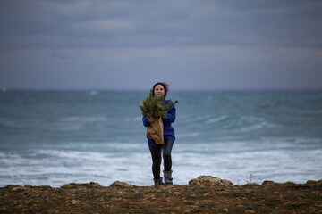 A beautiful girl with long red hair holds a craft bag with Christmas tree branches. A woman is dressed in a white sweater and a hat, she is walking along the winter coast of the sea.