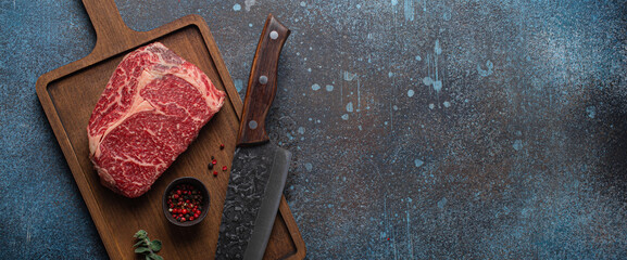 Raw meat beef marbled prime cut steak Ribeye on wooden cutting board and rustic concrete kitchen table background from above with big knife and spices, beefsteak concept