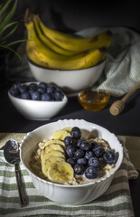 porcelain bowl with oatmeal, sliced ​​bananas and blueberries, decorated with honey and ingredients. Healthy breakfast still life background