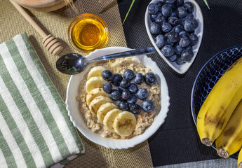 healthy breakfast of porcelain bowl with oatmeal, sliced ​​bananas and blueberries, decorated with honey and ingredients