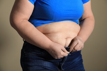 Overweight woman trying to button up tight jeans on light brown background, closeup