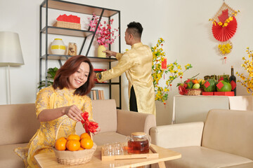 Smiling woman decorating basket of mandarins with red bow to present it as a gift for Tet to friends