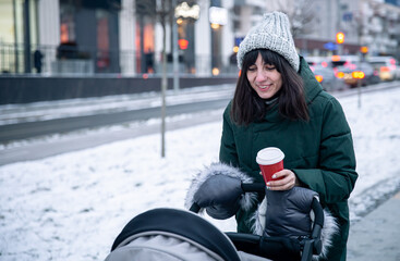 Stylish young mom with a cup of coffee on a walk with a baby carriage in winter.