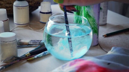 Close-up, the artist dilutes the paint with water, preparing to paint with paints. A woman's hand lowers an artistic brush into the water, the blue paint dissolves.