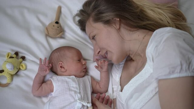 Happy woman talking with newborn boy lying on comfortable soft white bed at home. Top view portrait of excited Caucasian mother admiring infant baby kissing tiny hand in slow motion