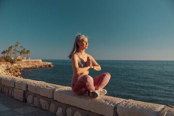 Fototapeta na wymiar Sporty woman in sports clothes sitting by the beautiful seashore in lotus position and taining on warm day