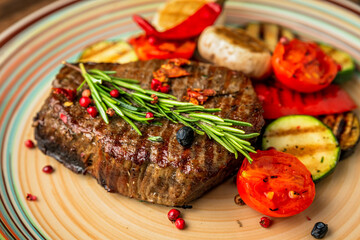 A close-up of a beef steak with grilled vegetables garnish, zucchini, aubergine, mushroom, pepper, rosemary, cherry tomato
