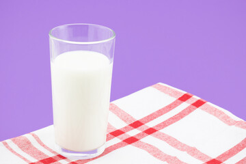 Full glass of milk on a red and white checkered linen napkin on a purple background and copy space. Healthy diet and source of calcium