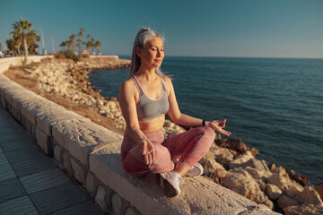 Fototapeta na wymiar Lady in sports clothes sitting by the beautiful seashore in lotus position and taining on warm day