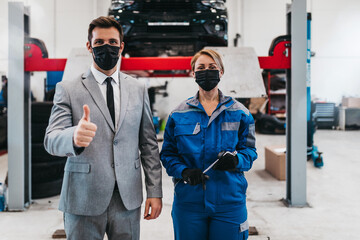 A businessman in a modern car repair shop is waiting for a mechanic to fix his car. Mechanic and client are wearing protective face masks due to Covid-19 pandemic.