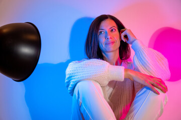 Smiling young brunette woman wearing a knitted sweater and white pants lit with a blue spotlight and a pink spotlight