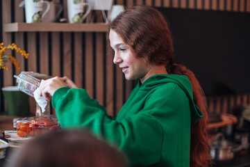 Side view of a young teen girl with red hair wearing bright green hooded sweatshirt cooking standing by table in living room