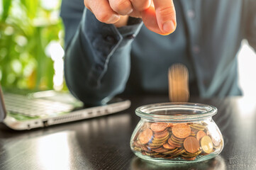 Mann schmeißt Geld in ein Glas voller Münzen auf einem Schreibtisch mit Laptop