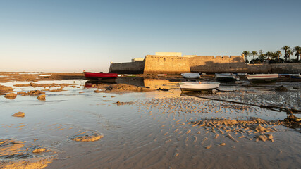 Boats docked in La Caleta beach at low tide with Santa Catalina Castle in the background at sunrise, Cadiz, Andalusia, Spain