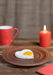 A fried egg in the shape of a heart lies on a clay plate with a red mug and a burning candle on a wooden table in a rustic style. Selective focus.