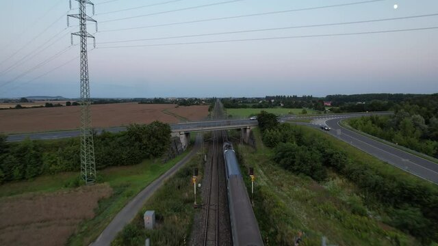 Aerial panorama of a passenger train passing under a road bridge in a rural area, surrounded by fields and trees.

