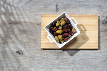 Deep square porcelain bowl with green and dark olives on a wooden board with shadow from a palm tree on a blue background